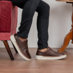 Imagem de um homem sentado, mostrando apenas do joelho para baixo, dando destaque para o sapatênis de couro legítimo em seus pés.